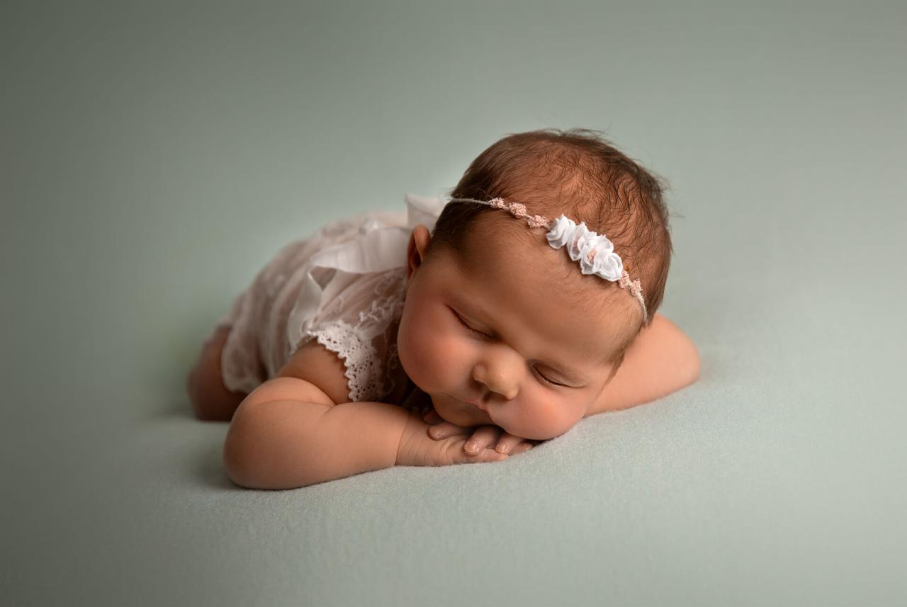 Śpiąca noworodka w koronkowej sukience i opasce z białymi kwiatami na delikatnym, pistacjowym tle.