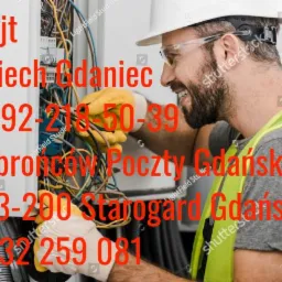 Elektryk w kasku i okularach ochronnych, w trakcie pracy przy skrzynce z przewodami, w żółtych rękawicach, uśmiechnięty, w tle kable i bezpieczniki.