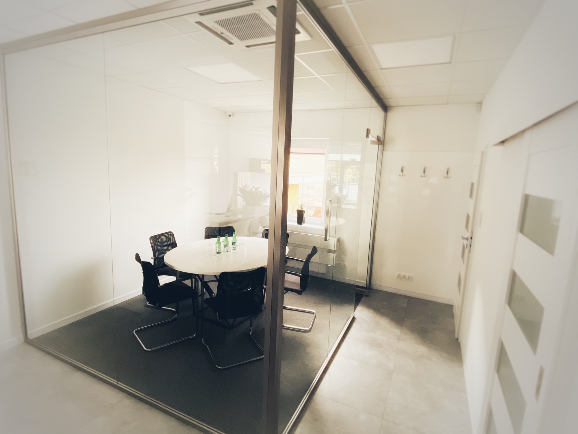 Nowoczesna sala konferencyjna za szklaną ścianą, okrągły stół z krzesłami, butelki wody i szklanki na stole, widok z korytarza, minimalistyczny design.