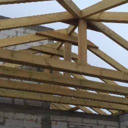 Konstrukcja drewniana dachu z widocznymi elementami więźby, mocowana do murowanych ścian budynku w trakcie budowy.