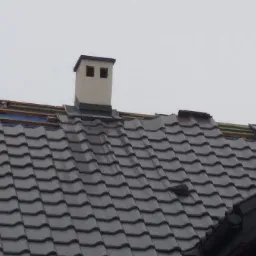 Fragment dachu z ciemną dachówką ceramiczną, widoczne dwa kominy, częściowo odkryta konstrukcja z łat i folii, ceglany fragment ściany szczytowej.