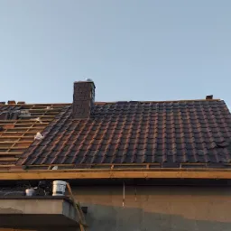 Częściowo pokryty dach z brązową dachówką, widoczne elementy więźby, komin z szarej dachówki i drugi komin w trakcie budowy z surowego drewna, rusztowanie z boku budynku.