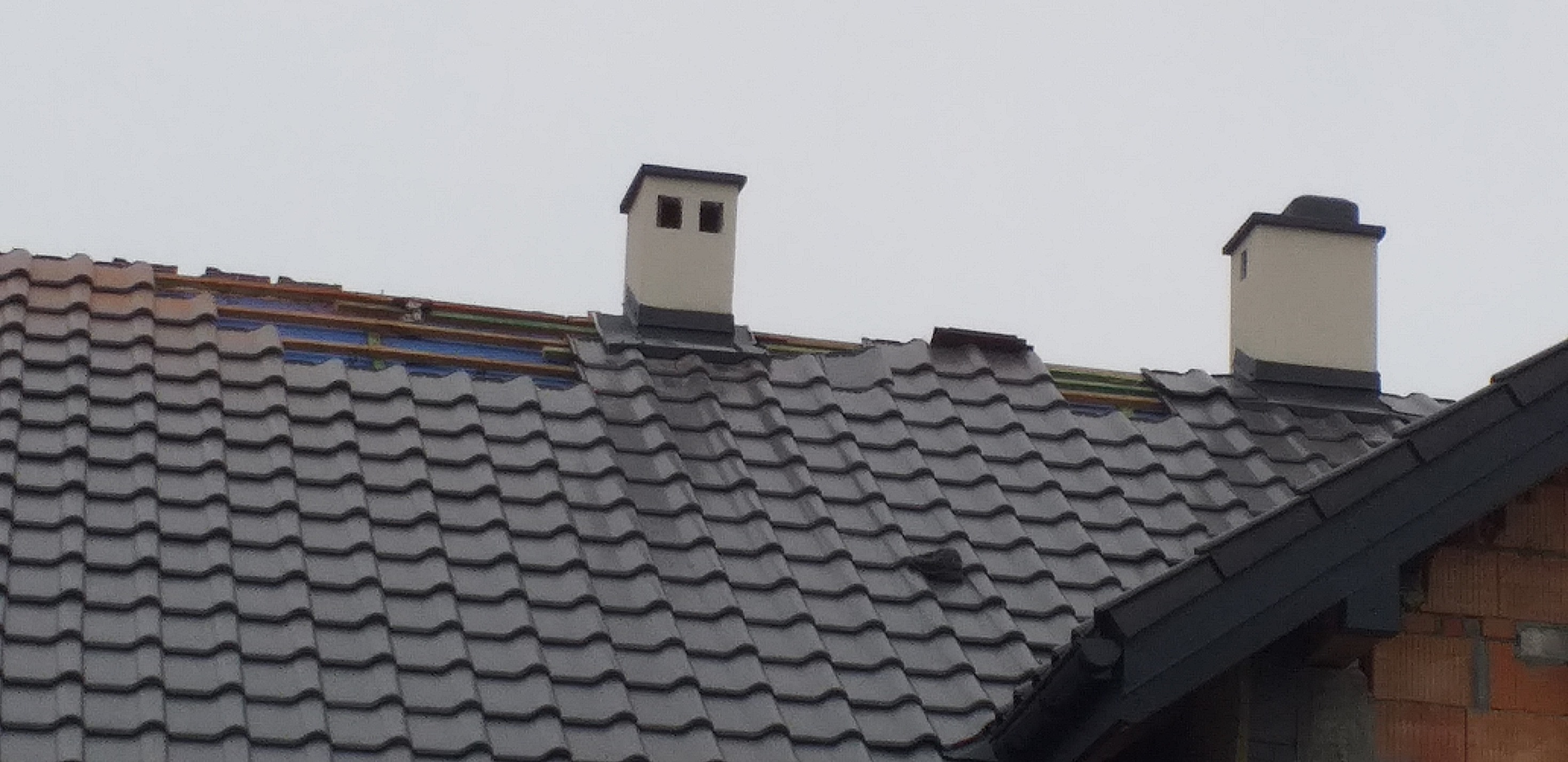 Fragment dachu z ciemną dachówką ceramiczną, widoczne dwa kominy, częściowo odkryta konstrukcja z łat i folii, ceglany fragment ściany szczytowej.