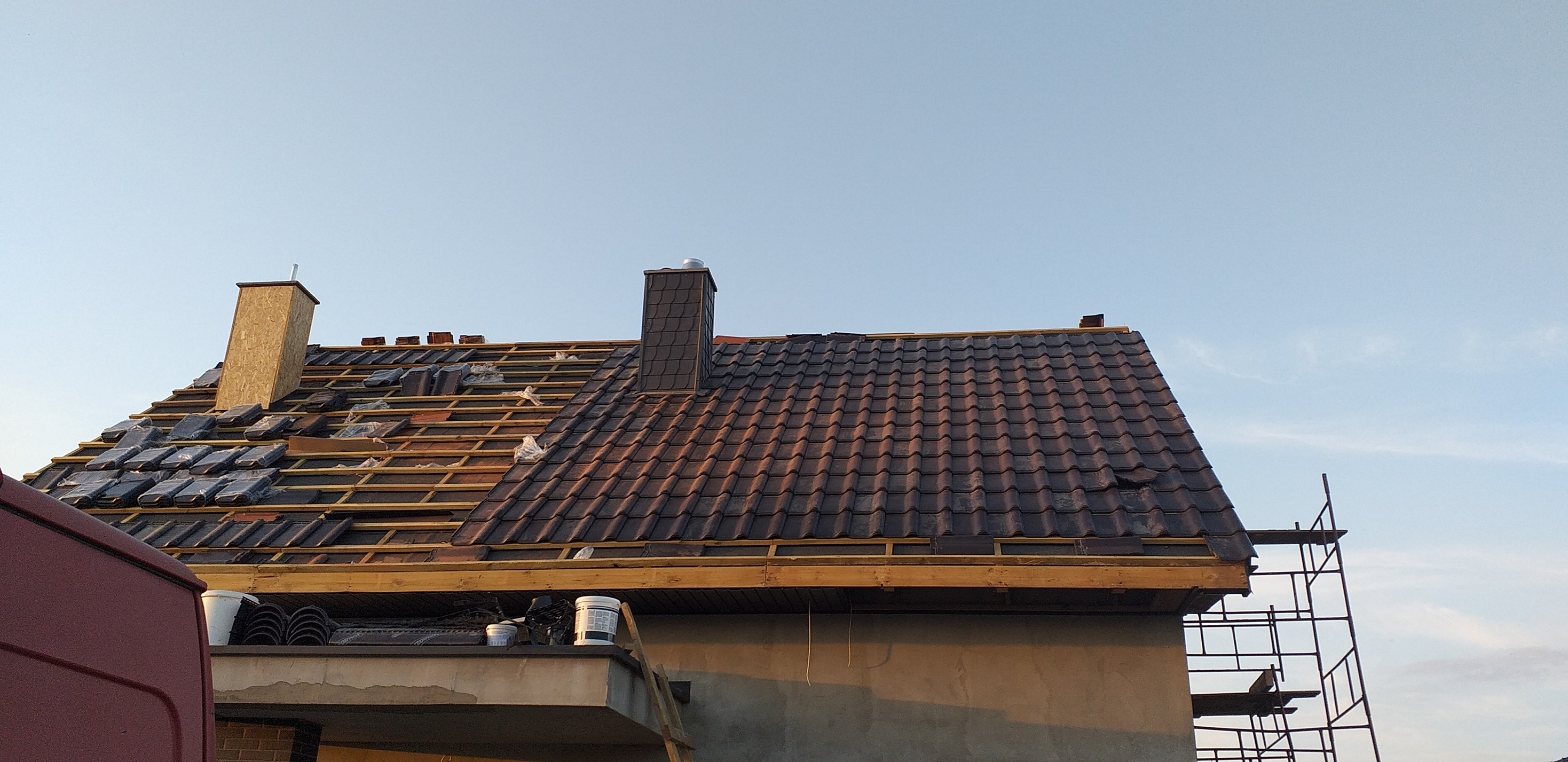 Częściowo pokryty dach z brązową dachówką, widoczne elementy więźby, komin z szarej dachówki i drugi komin w trakcie budowy z surowego drewna, rusztowanie z boku budynku.