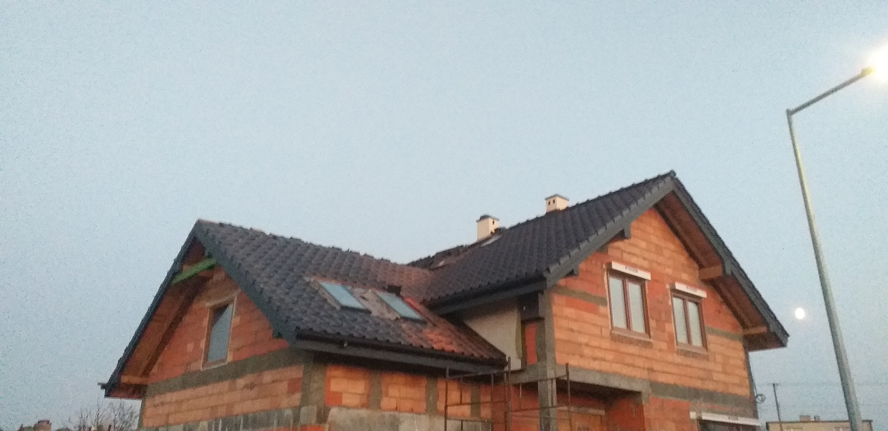 Niedokończony dom murowany z ciemną dachówką, widoczne okna dachowe i kominy, w tle słup oświetleniowy i pochmurne niebo.