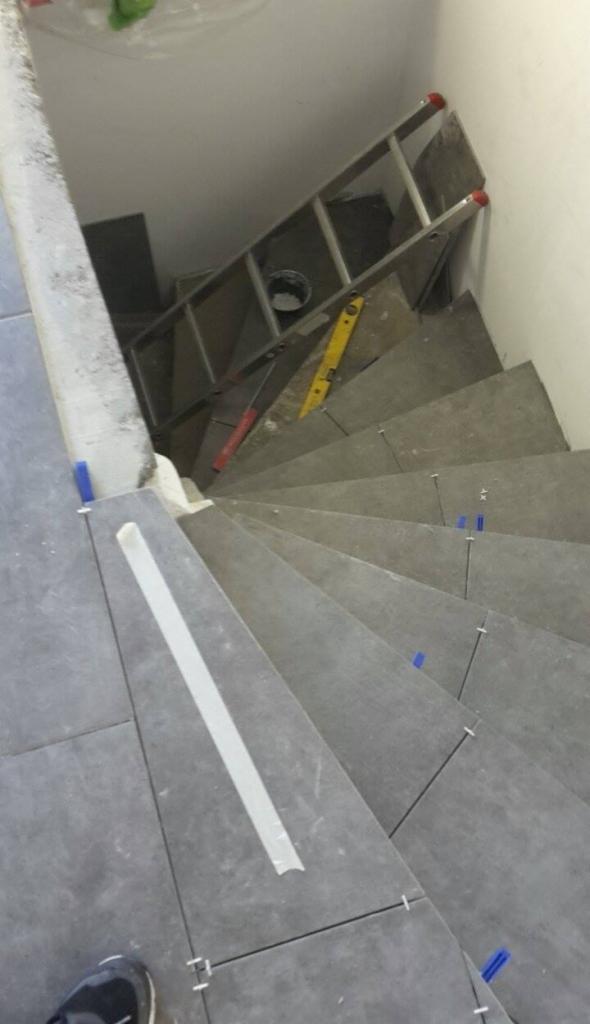 Układanie szarych płytek na schodach, widoczne poziomica, drabina i krzyżyki dystansowe, taśma malarska na krawędzi stopnia.