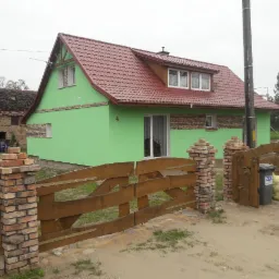 Dom jednorodzinny po remoncie elewacji, pomalowany na zielono z czerwonym dachem, ogrodzony drewnianym płotem z ceglanymi słupkami. Widoczny kosz na śmieci.