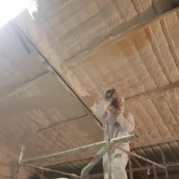 Pracownik w kombinezonie ochronnym aplikuje pianę izolacyjną na metalowy dach od wewnątrz, stojąc na rusztowaniu; widoczny wąż doprowadzający pianę i pojemnik z pianą.