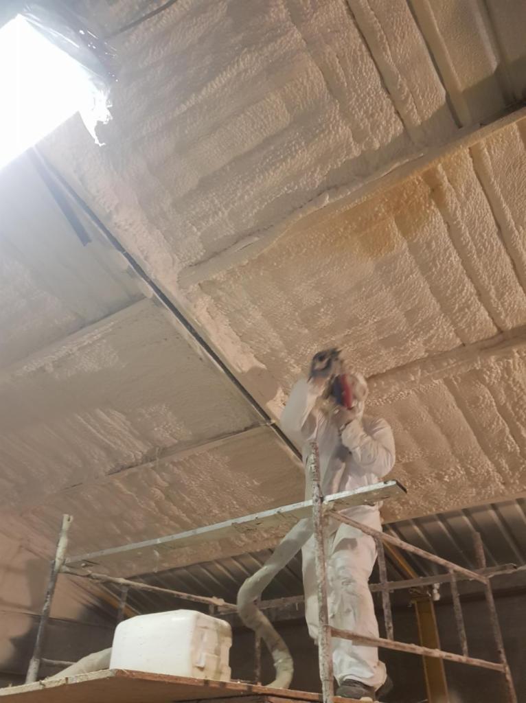 Pracownik w kombinezonie ochronnym aplikuje pianę izolacyjną na metalowy dach od wewnątrz, stojąc na rusztowaniu; widoczny wąż doprowadzający pianę i pojemnik z pianą.