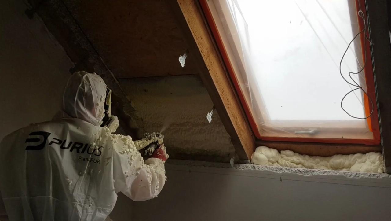Pracownik w kombinezonie ochronnym z logiem firmy PURIOUS natryskuje pianę izolacyjną na poddasze w pobliżu okna dachowego zabezpieczonego folią.