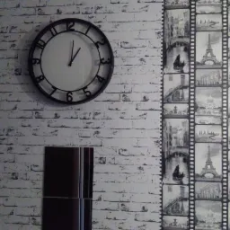 Ściana z tapetą imitującą białą cegłę, na której wisi czarny zegar z białym cyferblatem i ciemna, wąska szafka; obok fragment tapety z motywem taśmy filmowej przedstawiającej europejskie miasta.