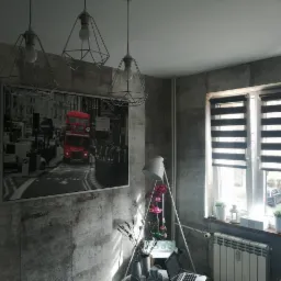 Pokój z panelami ściennymi imitującymi beton, wiszącą czarno-białą grafiką z czerwonym akcentem autobusu, trzema lampami drucianymi i oknem z żaluzjami dzień-noc.