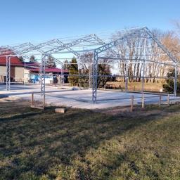 Haltech Group S.C. - Montaż szkieletu stalowego hali na wylanym fundamencie, widoczne osoby pracujące przy konstrukcji, budynek z czerwonym dachem w tle.