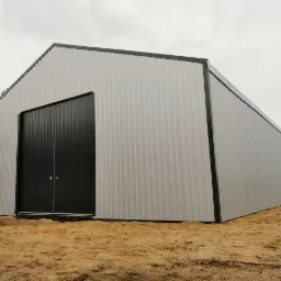 Gotowa hala z blachy trapezowej w kolorze srebrnym z czarnymi, dwuskrzydłowymi drzwiami na tle piaszczystego terenu i pochmurnego nieba.