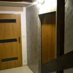 Wnętrze korytarza z drewnianymi drzwiami z przeszkleniami i ścianą pokrytą dekoracyjnym tynkiem imitującym kamień, oświetlone g&oacute;rnym światłem.