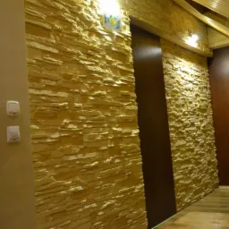 Wnętrze z dekoracyjną ścianą z kamienia piaskowego, brązowe drzwi przesuwne i drewniana podłoga, oświetlenie punktowe na suficie.