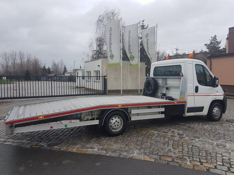 Biała laweta z platformą załadowczą, zaparkowana na brukowanej nawierzchni, widoczne reklamy firmy na flagach w tle.