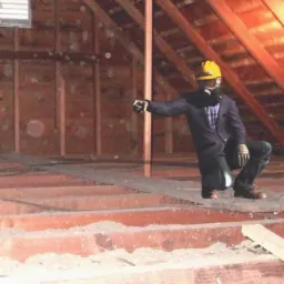 Inspektor w masce i kasku, w formalnym stroju, kuca w surowym stanie poddasza, wskazując na drewnianą konstrukcję dachu i ściany z desek.