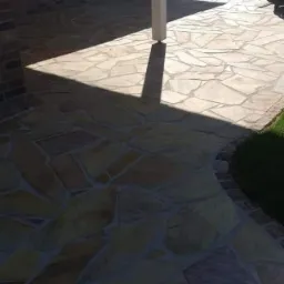 Ułożona z nieregularnych kamieni podłoga tarasu z widocznym cieniem słupa podtrzymującego zadaszenie, przylegająca do ściany budynku z cegły i trawnika.