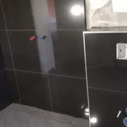 Częściowo wykończona łazienka z błyszczącymi czarnymi płytkami, widoczne przyłącza wodne i puszka elektryczna bez osprzętu, surowy fragment ściany w górnym rogu.