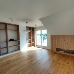 Manufaktura Nowych Wnętrz Paweł Nowak - Salon po wykończeniu: drewniana podłoga, zabudowy meblowe z półkami i szafkami z drewna, okno balkonowe w skosie. Jasne wnętrze.