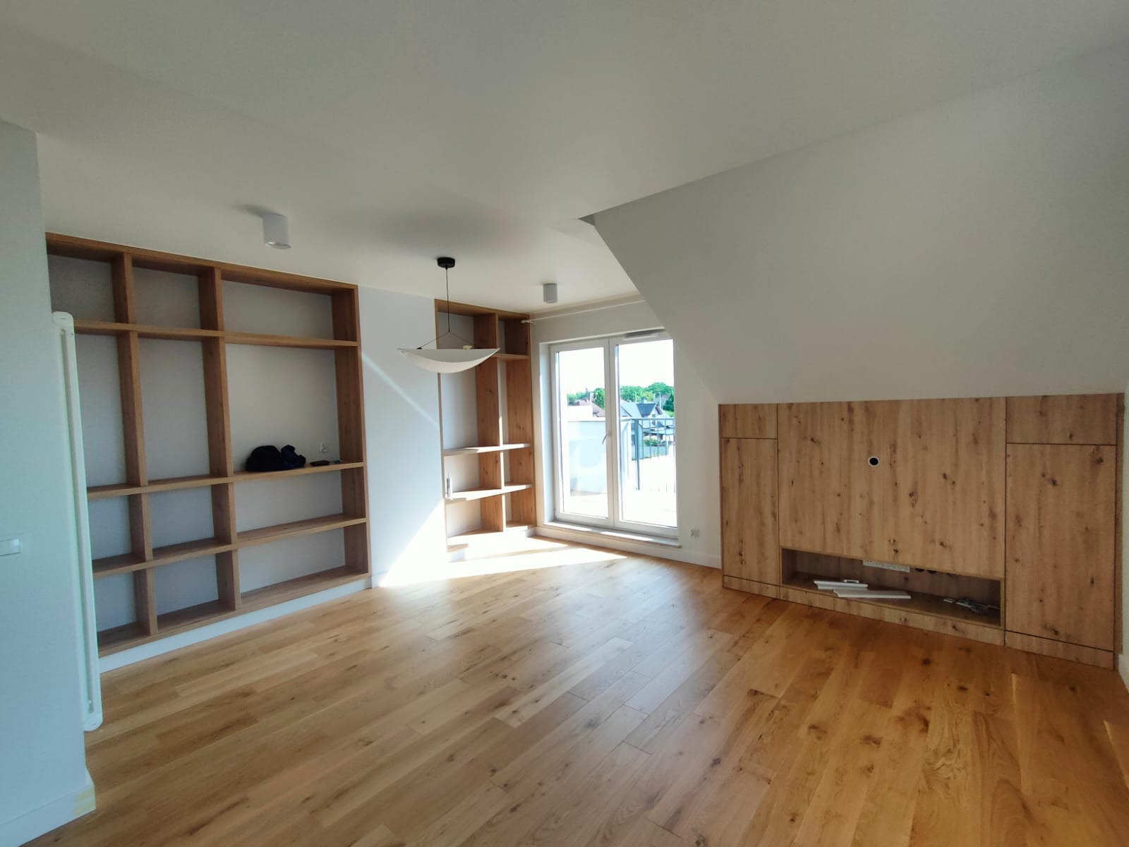 Salon po wykończeniu: drewniana podłoga, zabudowy meblowe z półkami i szafkami z drewna, okno balkonowe w skosie. Jasne wnętrze.