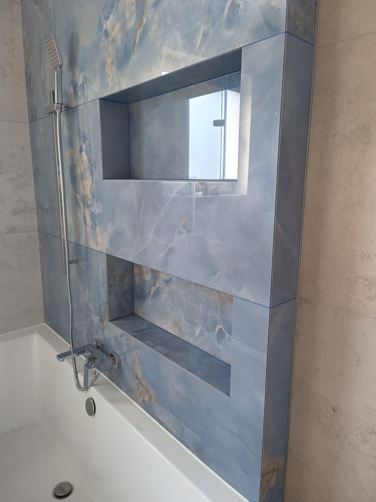 Niebieska, marmurkowa ściana nad wanną z wnękami, chromowana bateria i słuchawka prysznicowa. Wykończenie łazienki w nowoczesnym stylu.