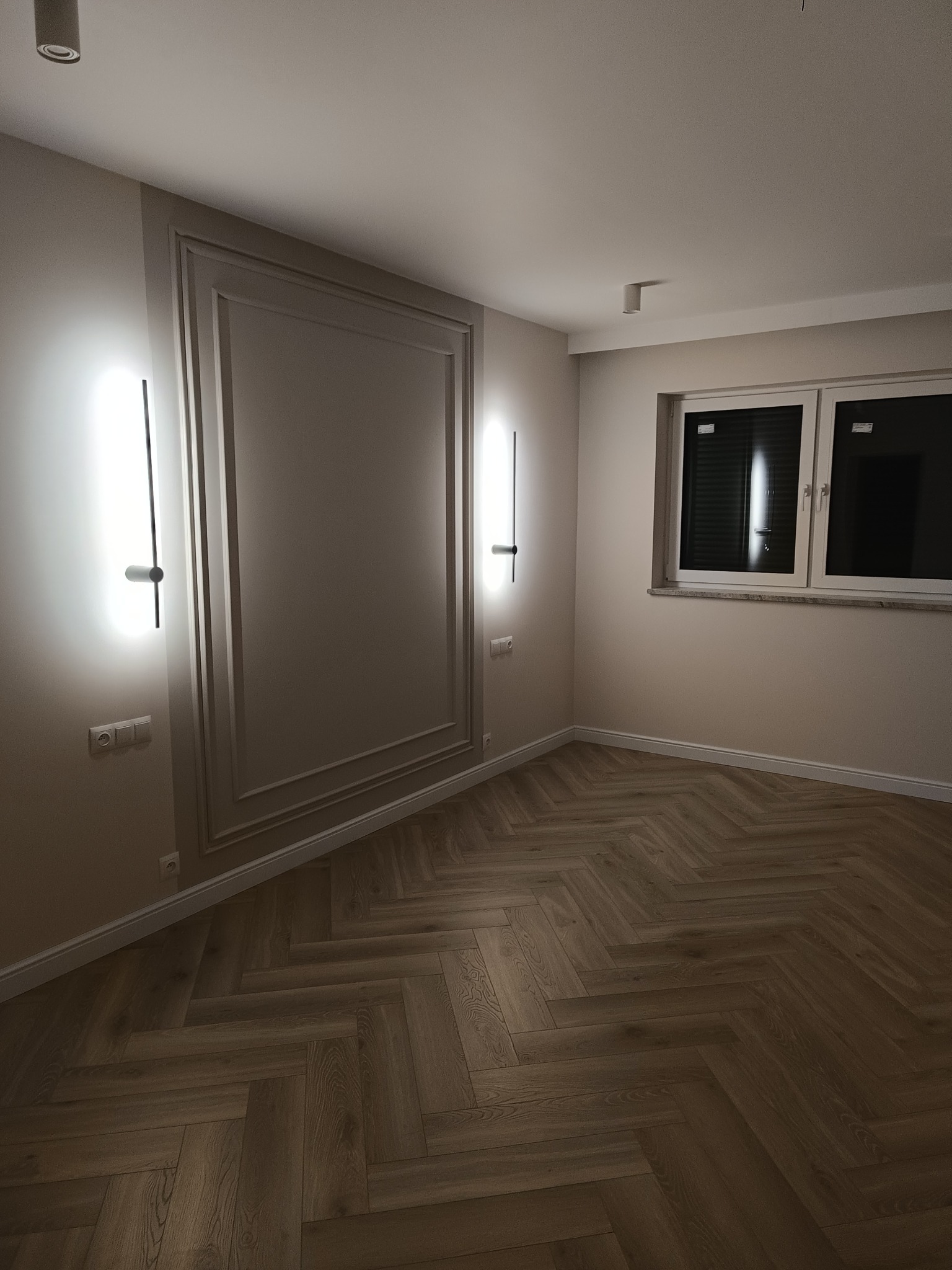 Jasna sypialnia po remoncie: parkiet w jodełkę, ozdobna rama na ścianie, nowoczesne kinkiety i okno z żaluzjami. Minimalistyczny design wnętrza.