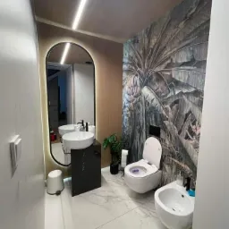 Nowoczesna łazienka z marmurową podłogą, umywalką na czarnym postumencie, toaletą i bidetem, z tapetą w motywy tropikalne i podświetlanym lustrem.