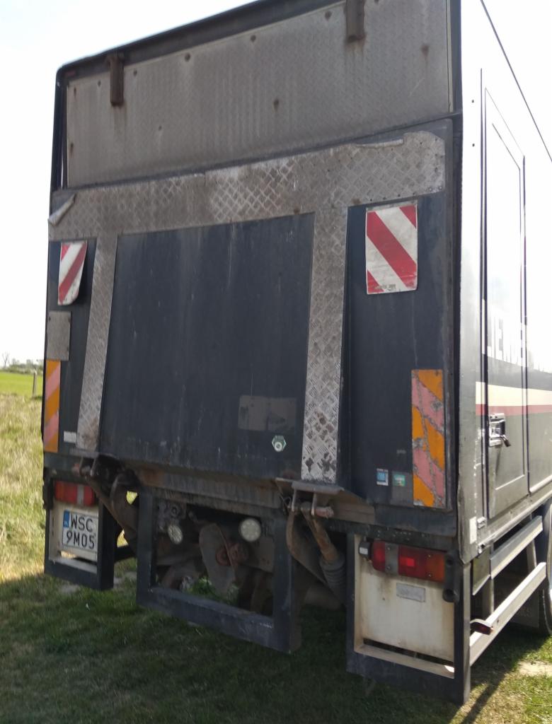 Tył brudnej, czarnej ciężarówki z podniesioną klapą, widoczne światła, tablica rejestracyjna i elementy konstrukcyjne na tle zielonej trawy i pola.