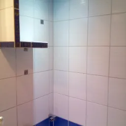 Wykończona ściana łazienki z białymi płytkami, dekoracyjnym pasem z motywem roślinnym i fragmentem baterii umywalkowej.