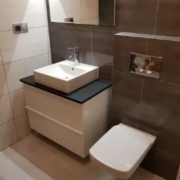 Nowoczesna łazienka z białymi płytkami i brązowymi akcentami, wisząca toaleta i kwadratowa umywalka nablatowa na białej szafce z czarnym blatem, lustro na ścianie.