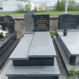 Szary, granitowy nagrobek z ciemniejszą tablicą zawierającą inskrypcje, krzyż i daty zmarłych, otoczony innymi nagrobkami na cmentarzu.