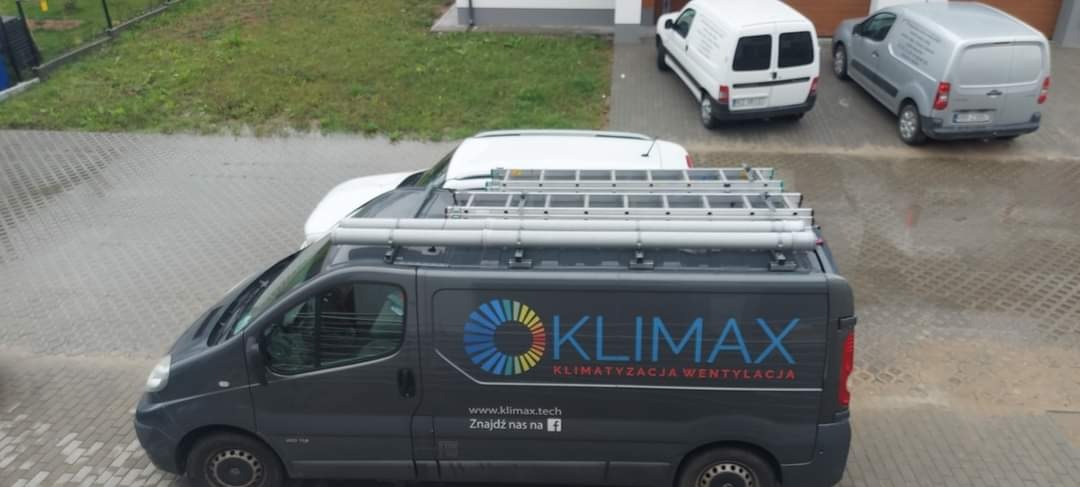 Szary samochód dostawczy z logo firmy Klimax Klimatyzacja Wentylacja, drabiny na dachu, zaparkowany na brukowanej powierzchni obok innych pojazdów w deszczowy dzień.