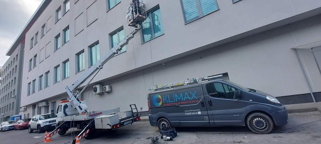 Pracownik na podnośniku koszowym przy elewacji biurowca, montaż lub serwis klimatyzacji zewnętrznej, obok zaparkowany samochód firmowy z logo.