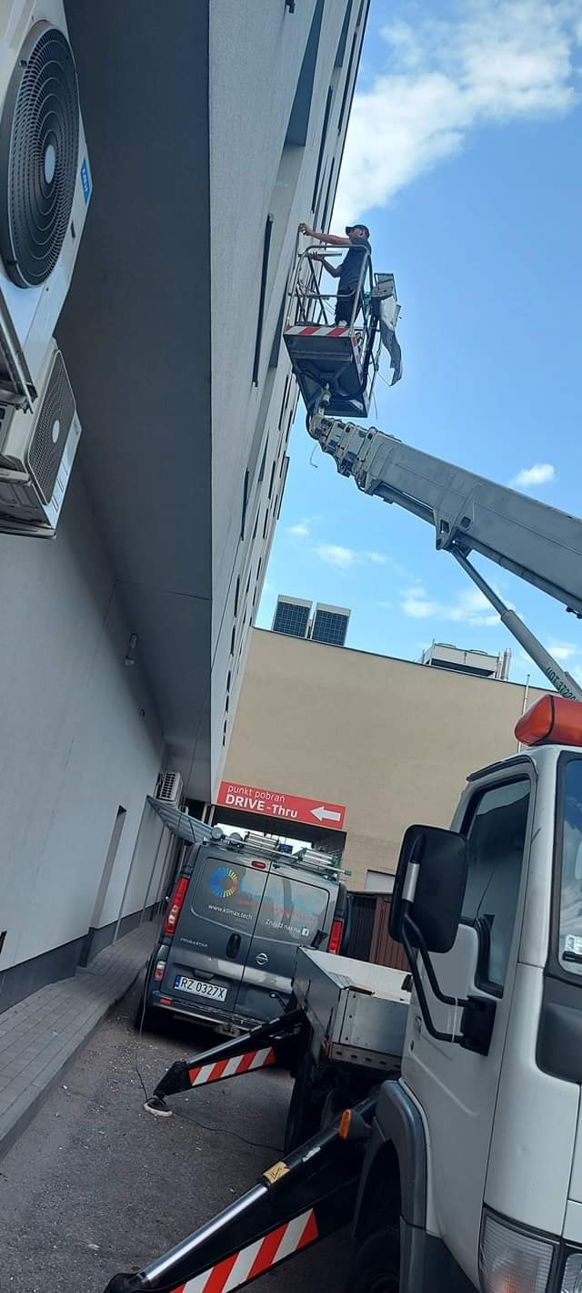 Pracownik na podnośniku koszowym montuje klimatyzator na elewacji budynku, widoczny firmowy van z logo i numerem rejestracyjnym oraz znak informacyjny punktu pobrań Drive-Thru.