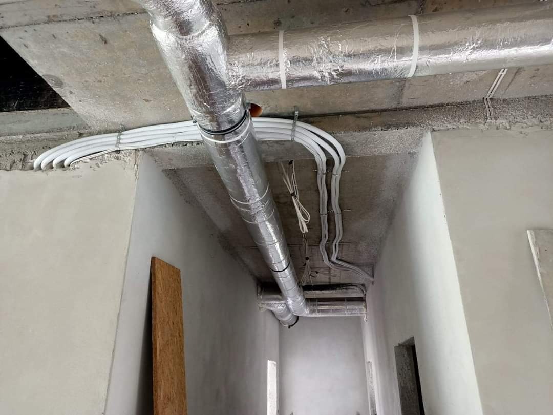 Montaż instalacji wentylacyjnej z rurami w srebrnej izolacji termicznej i wiązką białych kabli mocowanych do surowego, betonowego sufitu w surowym wnętrzu budynku.