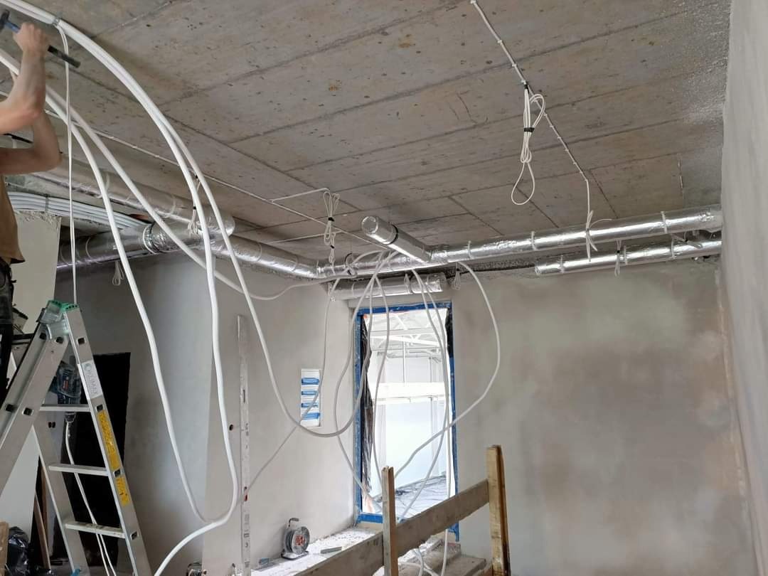 Montaż instalacji wentylacyjnej i przewodów elektrycznych w surowym wnętrzu biurowym, widoczne aluminiowe rury, białe przewody, drabina i częściowo widoczna osoba wykonująca prace.