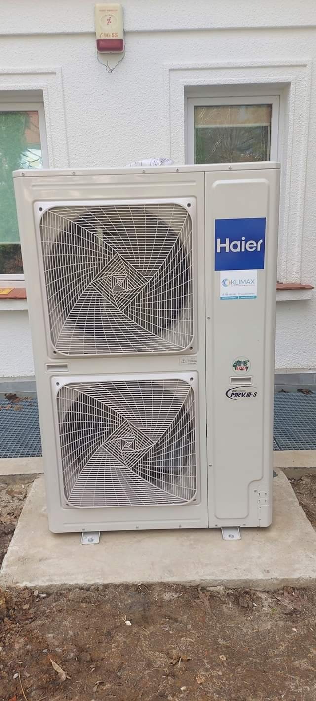 Zewnętrzna jednostka klimatyzacji Haier na betonowej podstawie przy budynku z białym tynkiem i dwoma oknami.