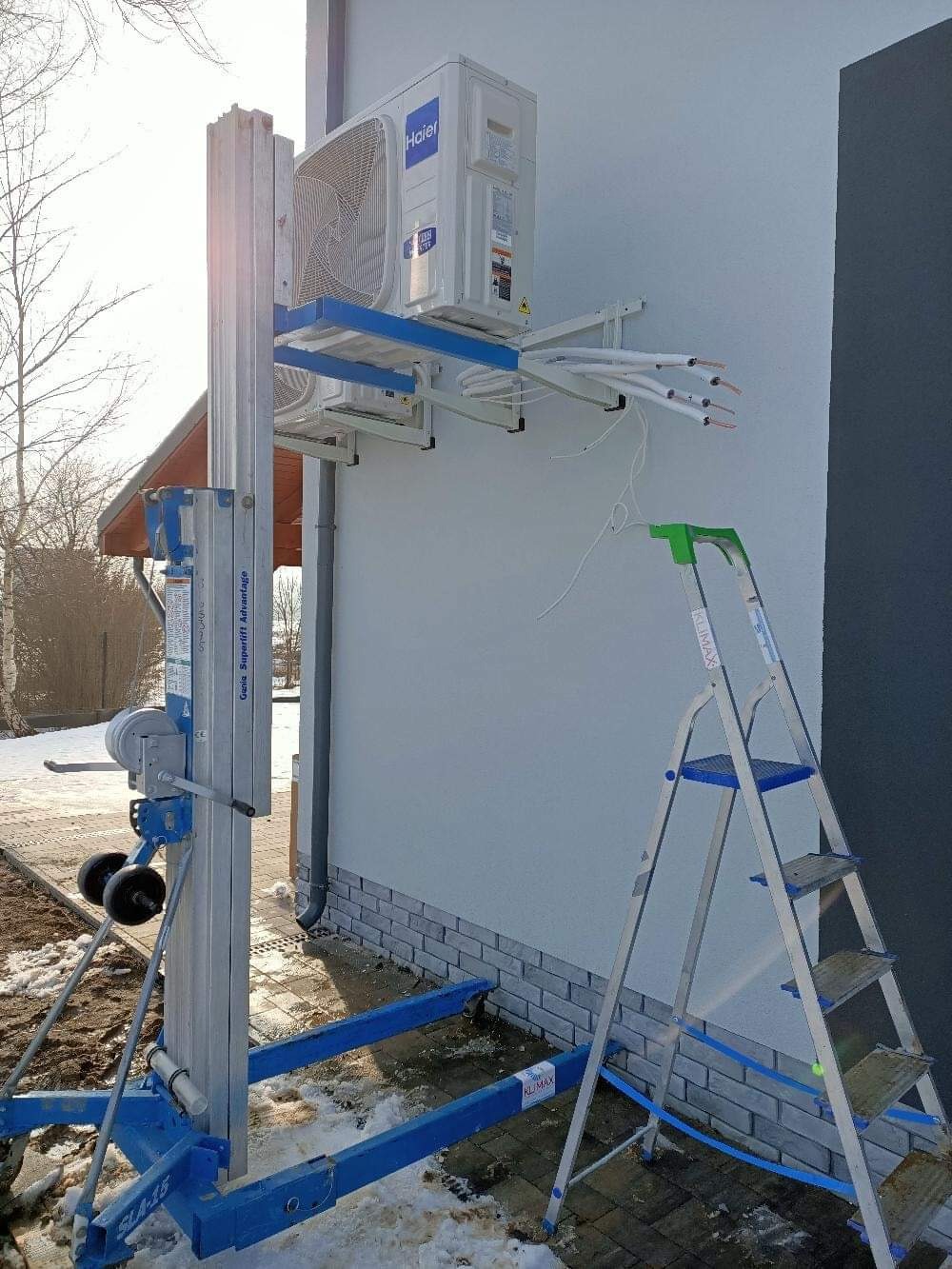 Montaż klimatyzatora Haier na elewacji budynku, widoczny podnośnik Genie Superlift Advantage i drabina Klimax, przewody instalacyjne wystają ze ściany.