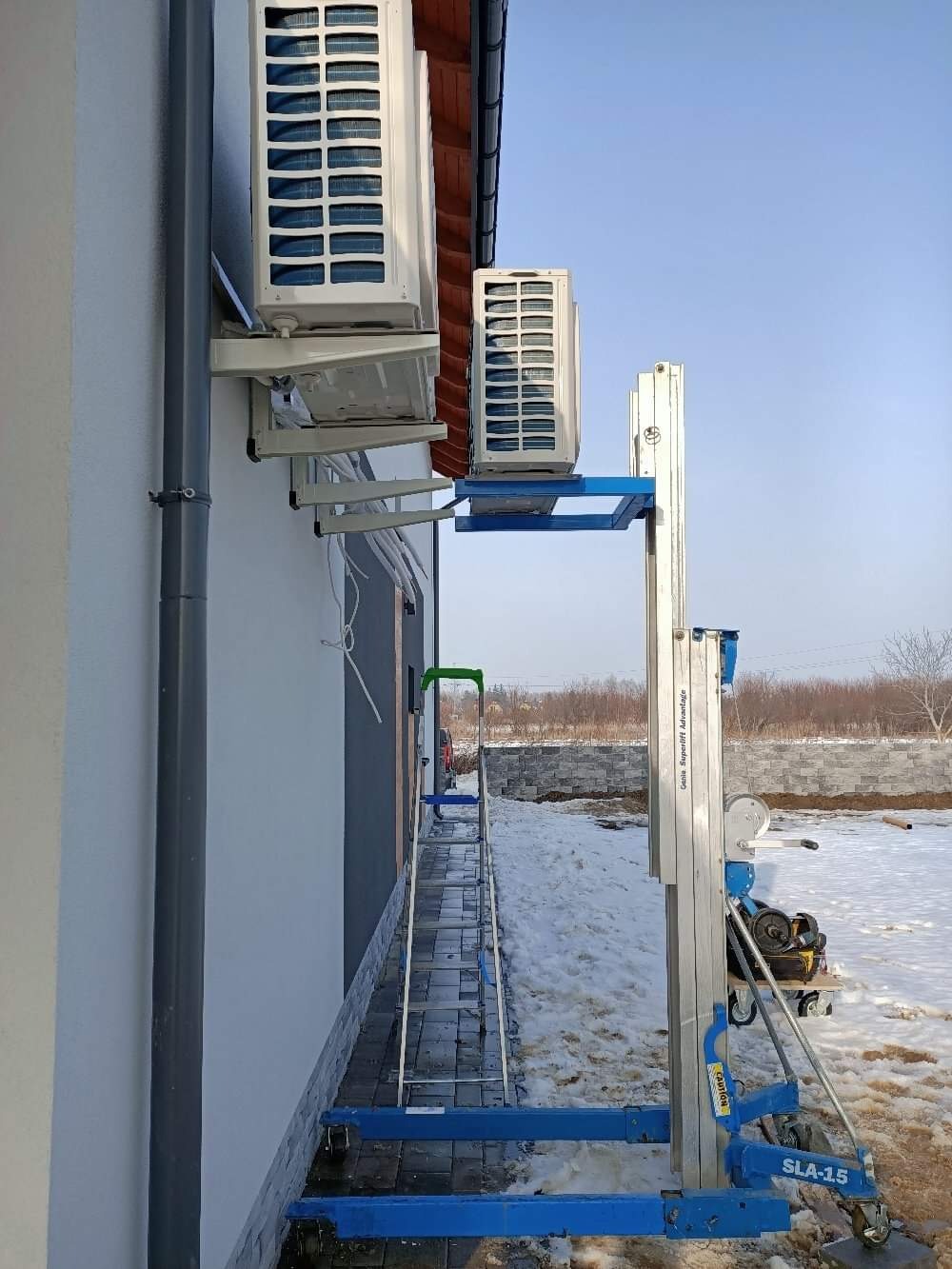 Dwa klimatyzatory zamontowane na ścianie domu, jeden nad drugim, z użyciem podnośnika teleskopowego Genie SLA-15 i drabiny, widoczne elementy instalacji.