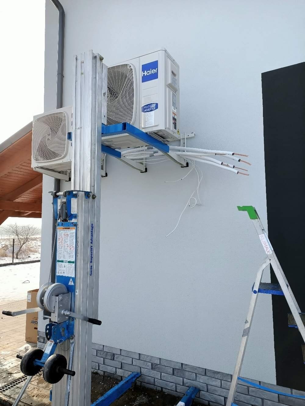 Montaż klimatyzacji: jednostka zewnętrzna marki Haier instalowana na ścianie budynku za pomocą podnośnika Genie Superlift Advantage, widoczne kable i drabina.