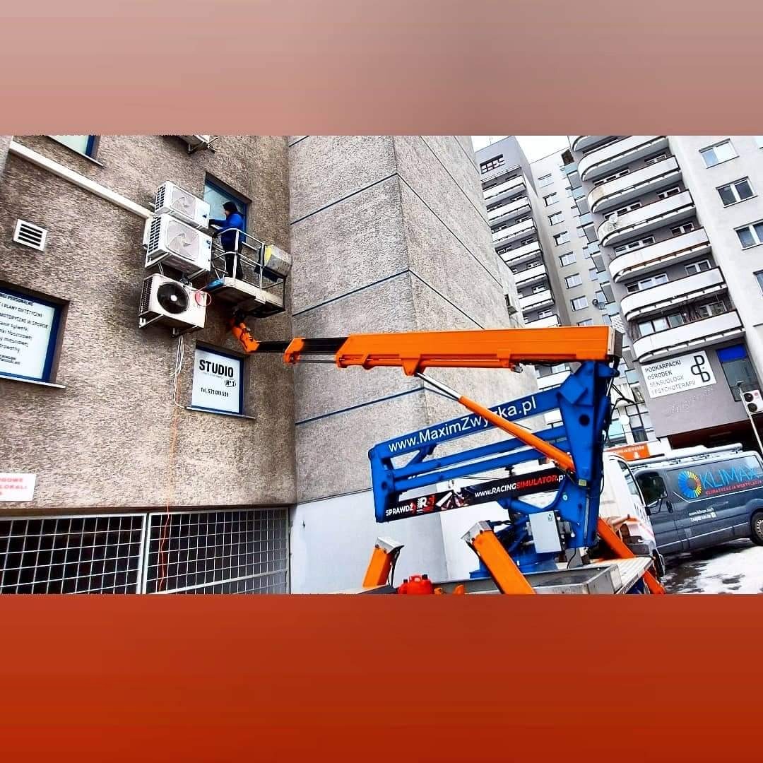 Montaż klimatyzacji na elewacji budynku z użyciem podnośnika koszowego, widoczna instalacja i pracownik w koszu. Na ścianie zamontowane trzy jednostki klimatyzacyjne.