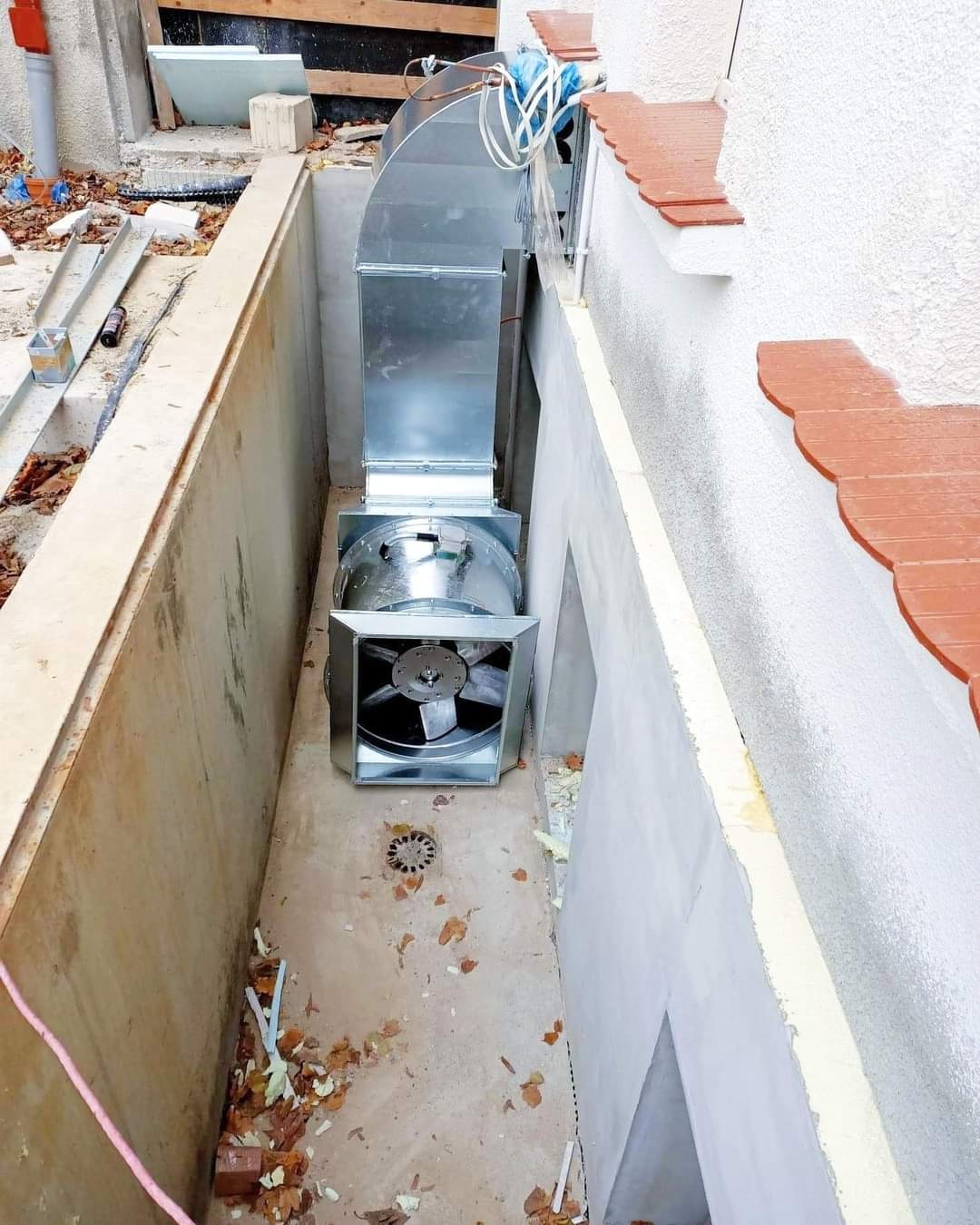 Montaż kanału wentylacyjnego z wentylatorem w betonowym szybie, widoczne liście i drobne odpady budowlane na dnie.
