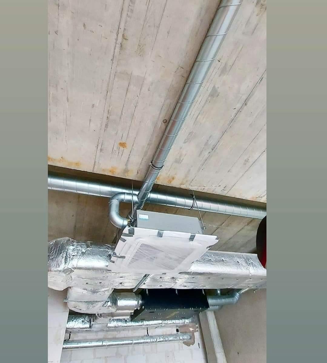 Widok instalacji klimatyzacji z widocznymi rurami wentylacyjnymi, centralą klimatyzacyjną i izolacją termiczną na surowym betonowym suficie.