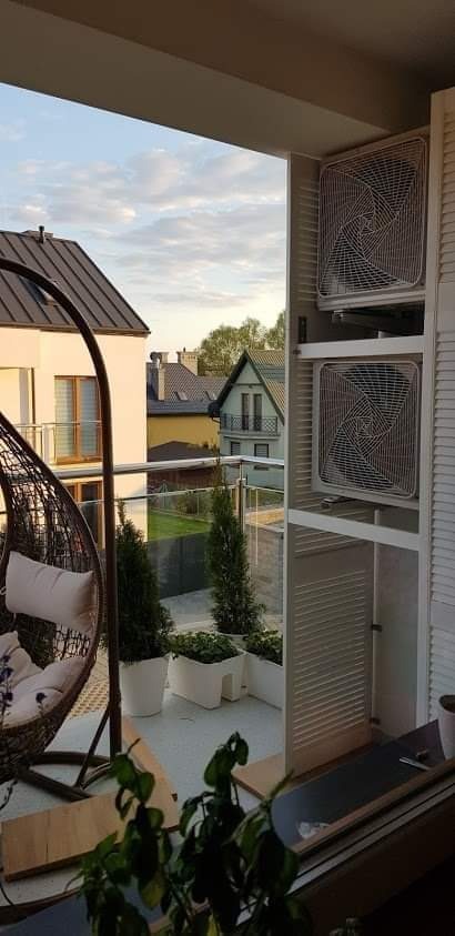 Dwa klimatyzatory zamontowane na balkonie w białej obudowie z żaluzjami, widok na osiedle domów jednorodzinnych i wiszące krzesło.