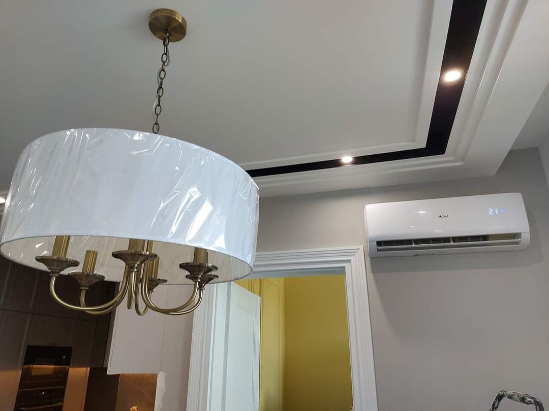 Biała klimatyzacja Haier zamontowana na szarej ścianie w pomieszczeniu z klasycznym wystrojem, widoczny żyrandol z abażurem w folii ochronnej i dekoracyjny sufit z oświetleniem punktowym.