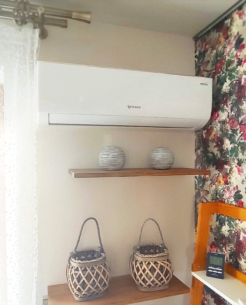 Biały klimatyzator Rotenso zamontowany w pokoju z tapetą w kwiaty, pod nim drewniane półki z dekoracjami, w tle firanka.