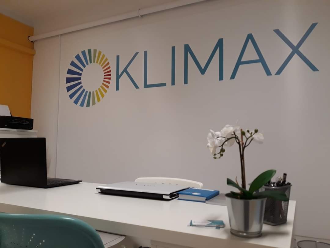 Biuro firmy z logo 'KLIMAX' na ścianie, biurko z laptopem, drukarką i doniczką z kwiatami.