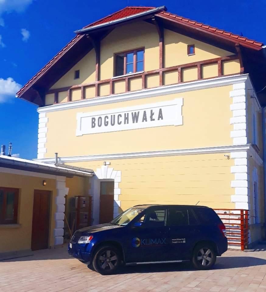 Żółty budynek stacji kolejowej Boguchwała z czerwonym dachem i niebieski samochód z logo Klimax na pierwszym planie.